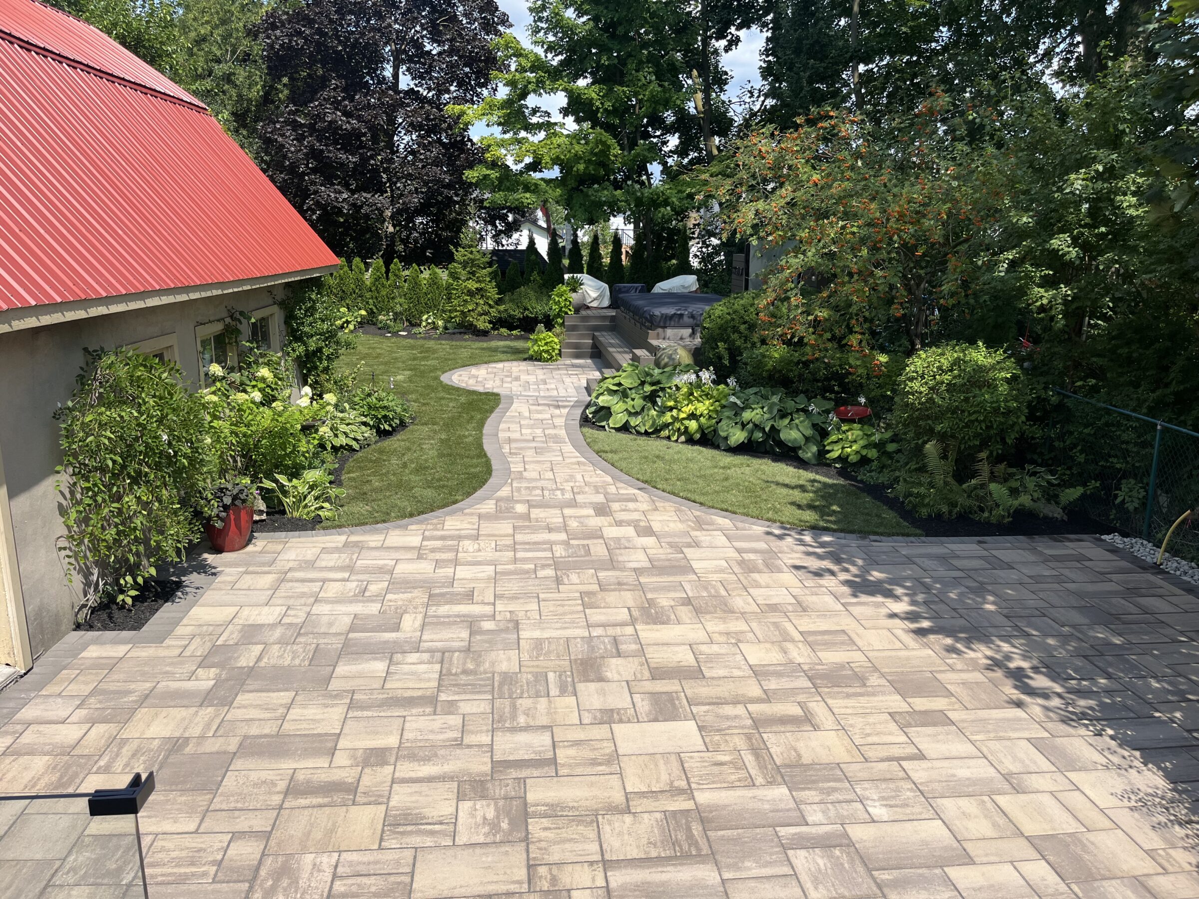 A red-roofed building and curving stone pathway through a lush garden with shrubs and trees under a sunny sky.