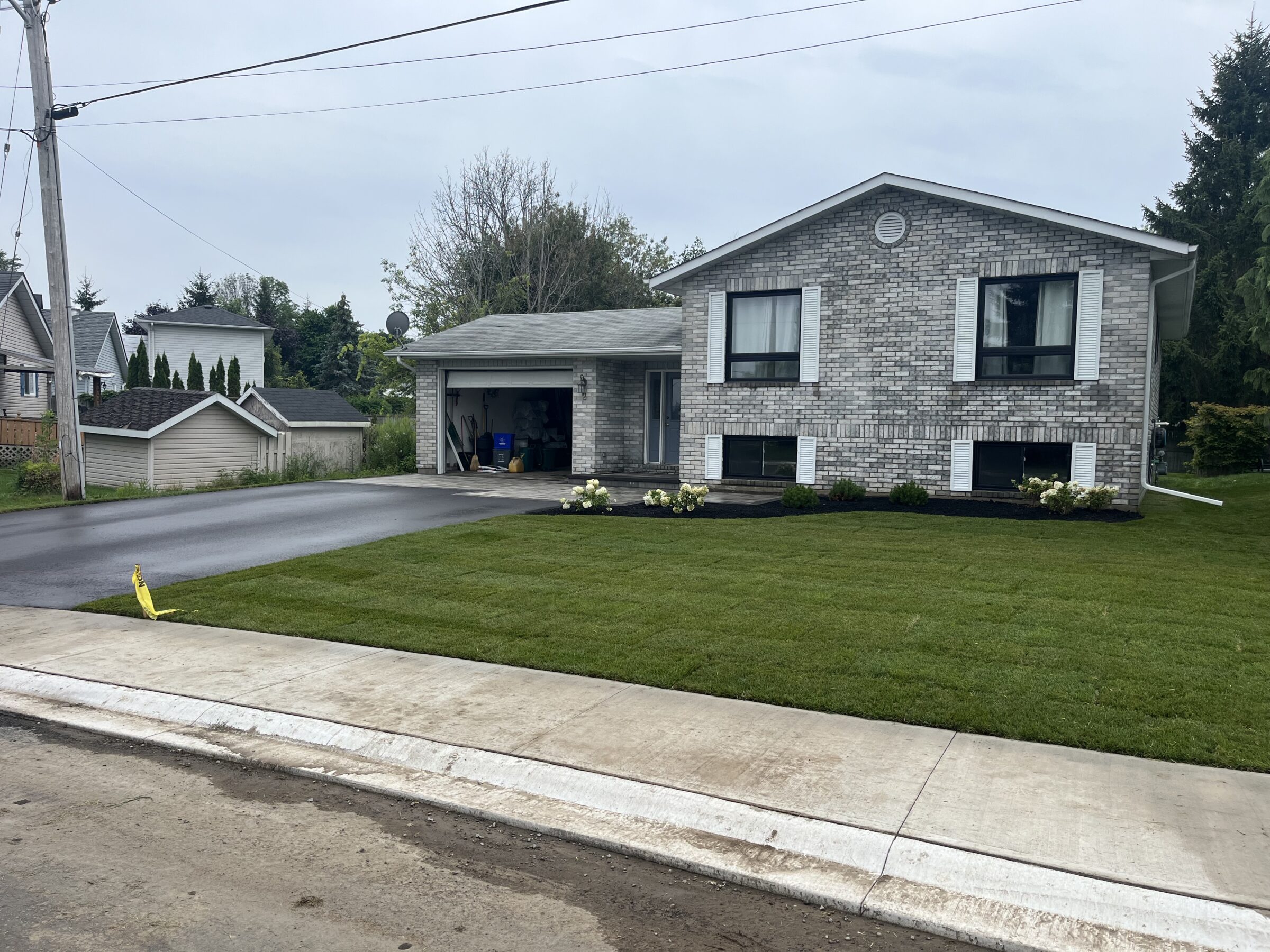 A grey brick house with a freshly landscaped yard and driveway, surrounded by trees and neighboring homes on a quiet suburban street.