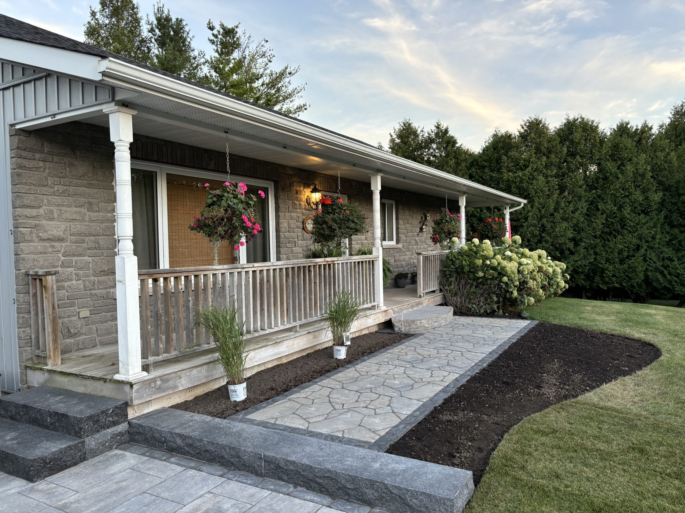 A stone house with a porch, hanging flowers, lush greenery, and a stone pathway set against a backdrop of trees under a cloudy sky.