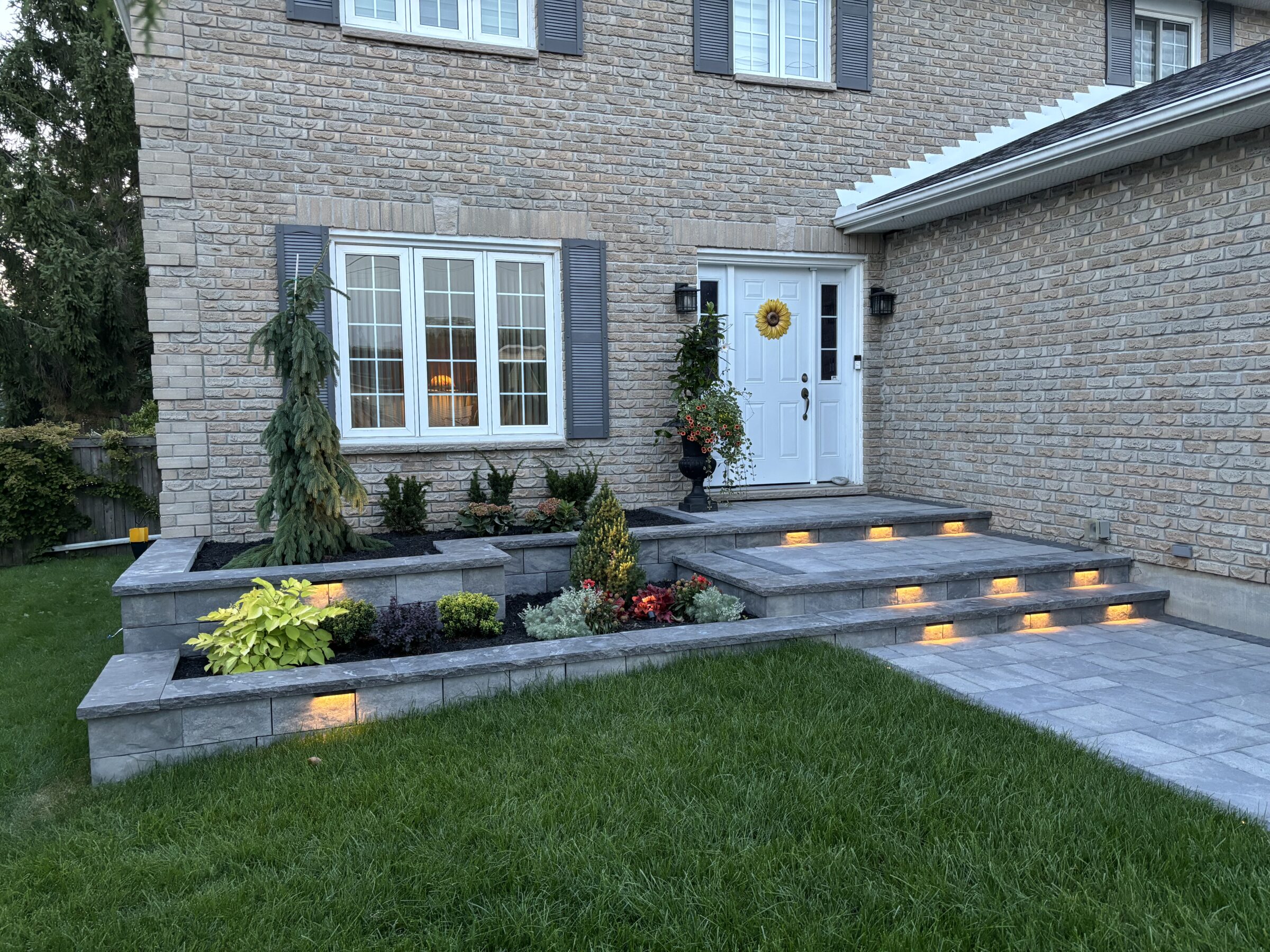 A brick house with a landscaped entrance features stone steps, illuminated by small lights, and a flower arrangement by the white door.