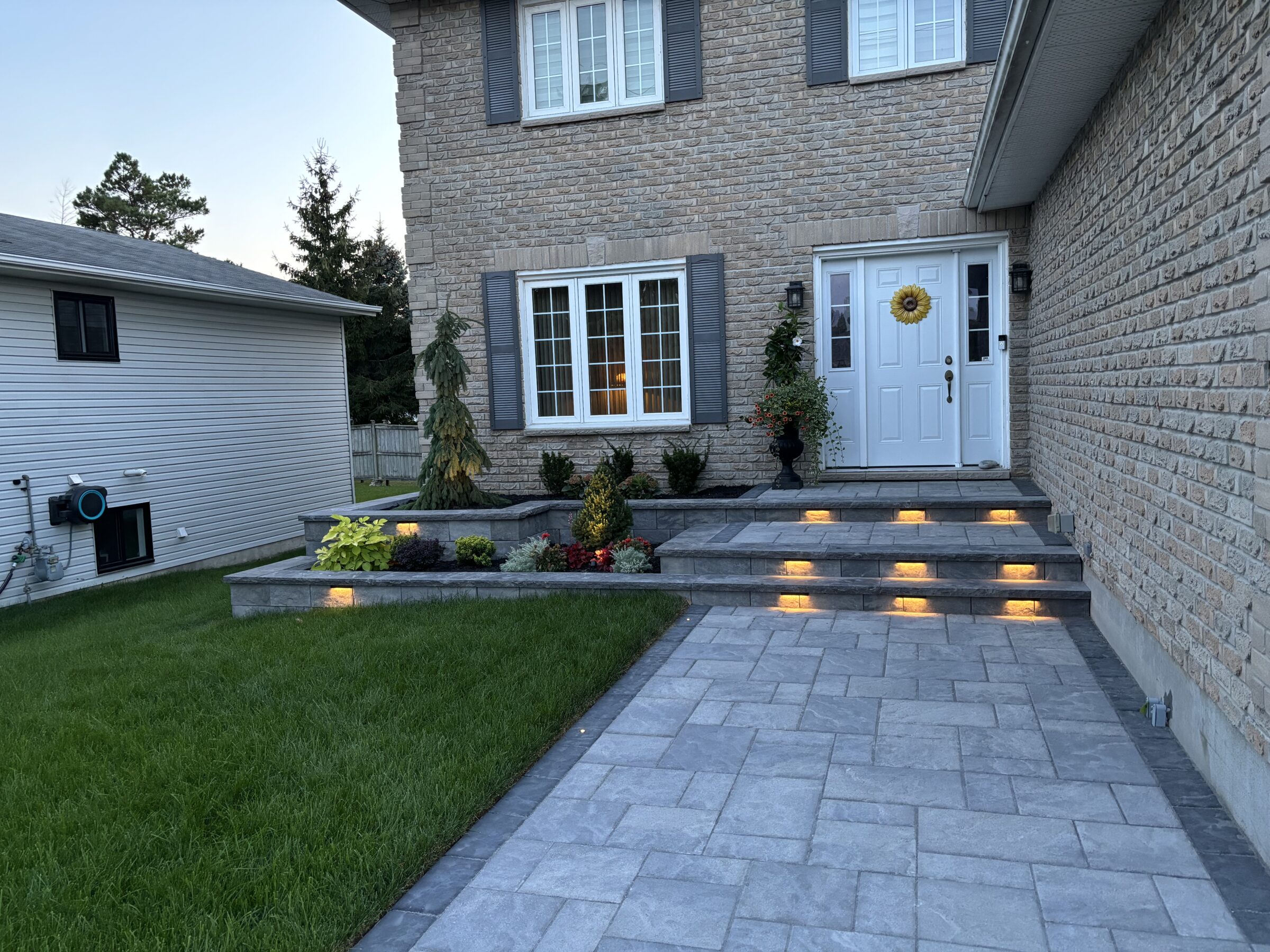 A brick house with a well-lit entrance and garden, featuring a pathway leading to a white door adorned with a sunflower wreath.