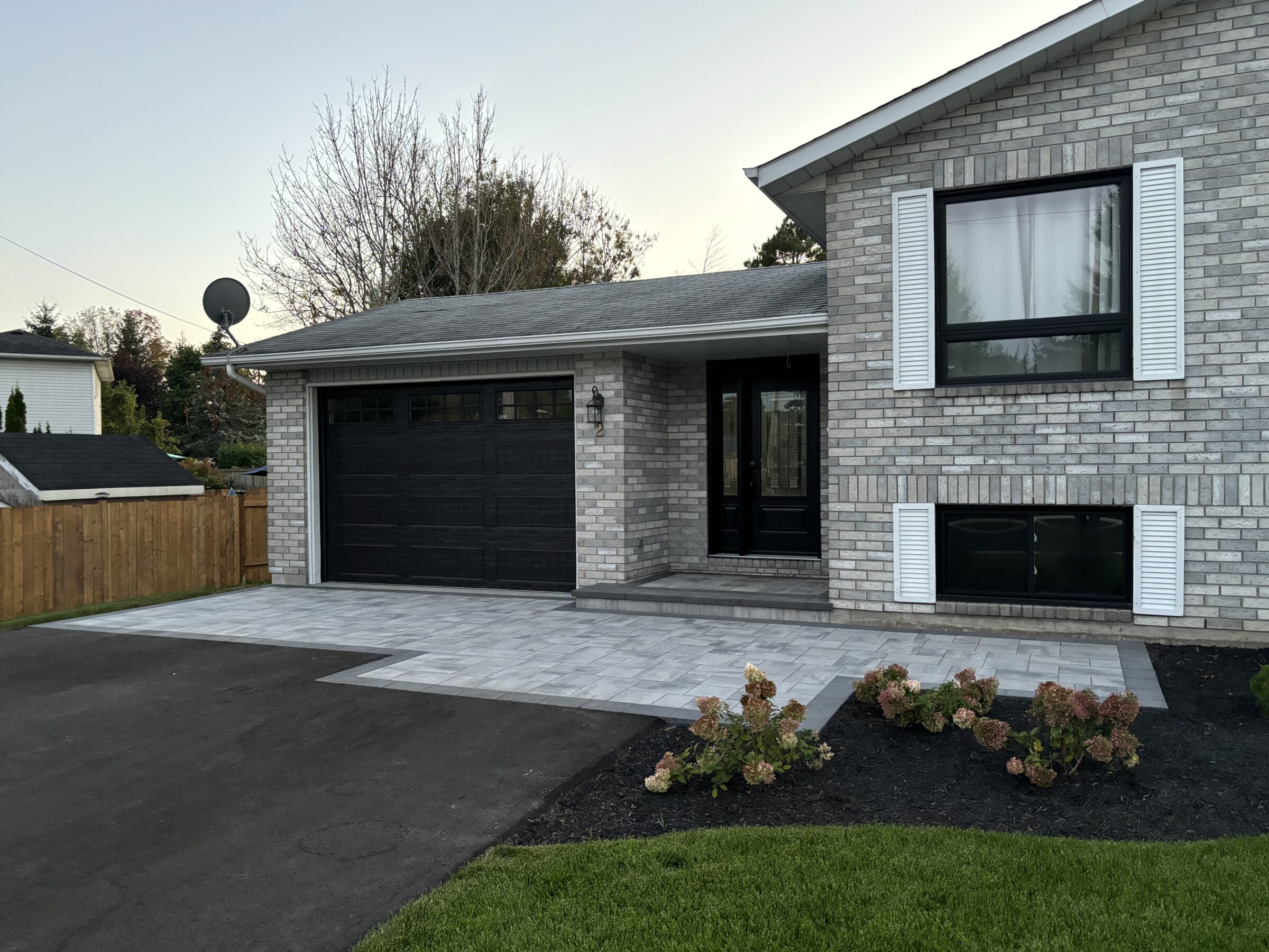 A modern brick house with black garage door, landscaped yard, patio, and surrounding trees. Calm suburban atmosphere with clear sky.