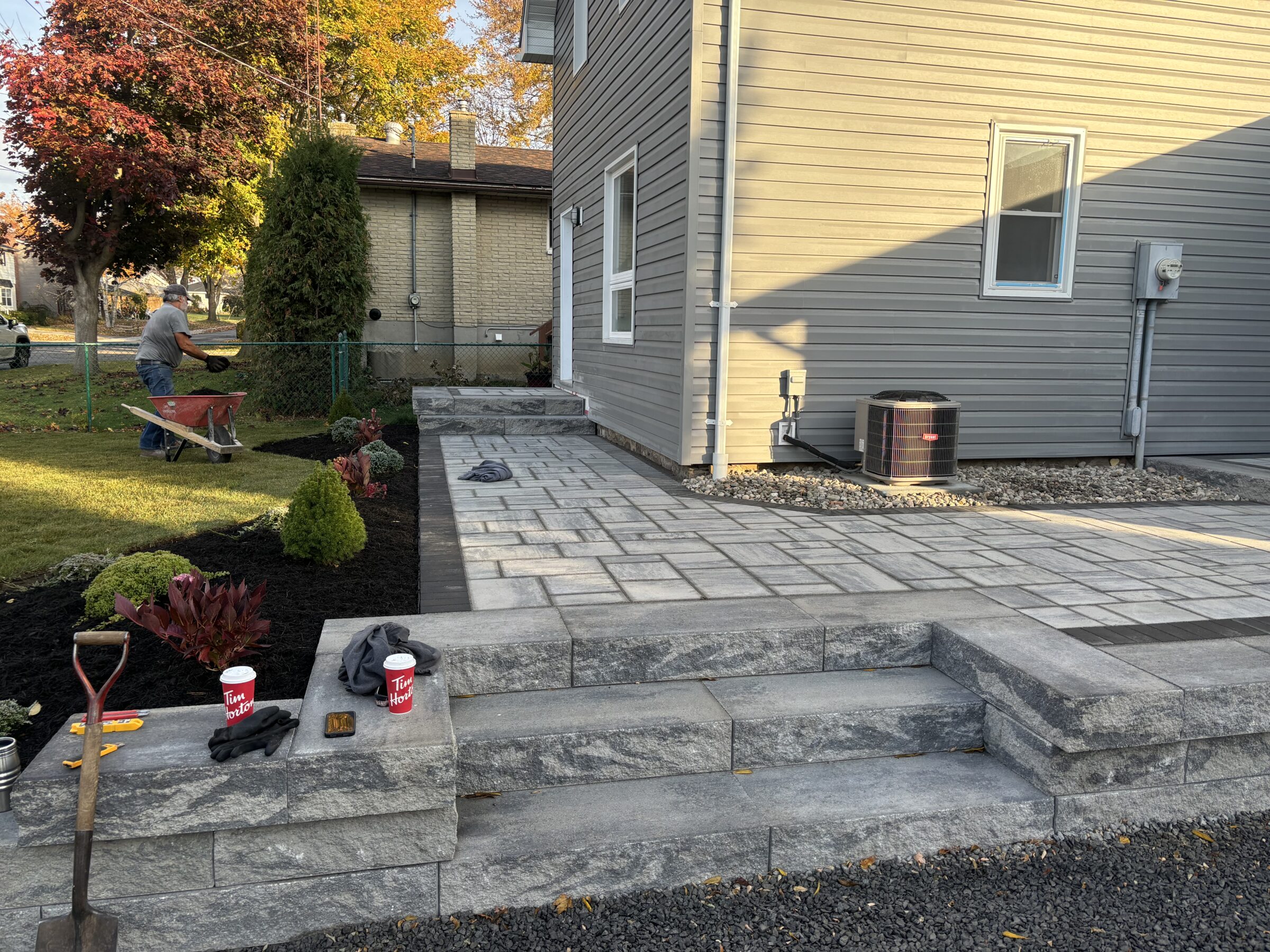 A person landscaping with a wheelbarrow near a paved patio beside a house; coffee cups on stone steps, autumn trees in background.