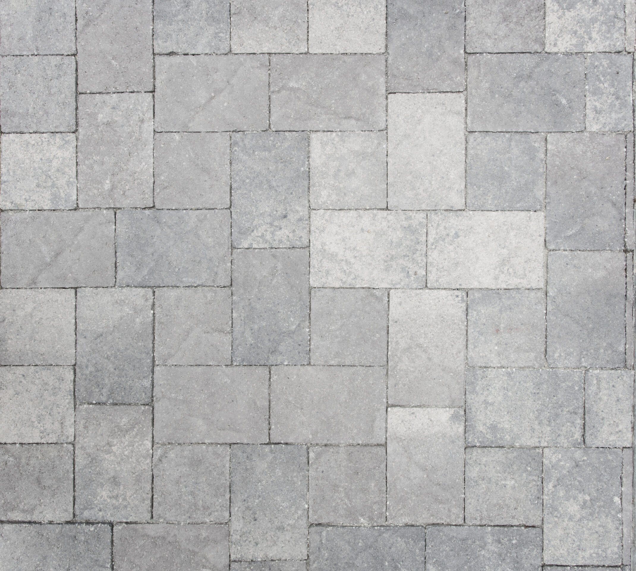 The image shows a close-up of gray, interlocking stone pavers arranged in a herringbone pattern, creating a textured surface with subtle color variations.