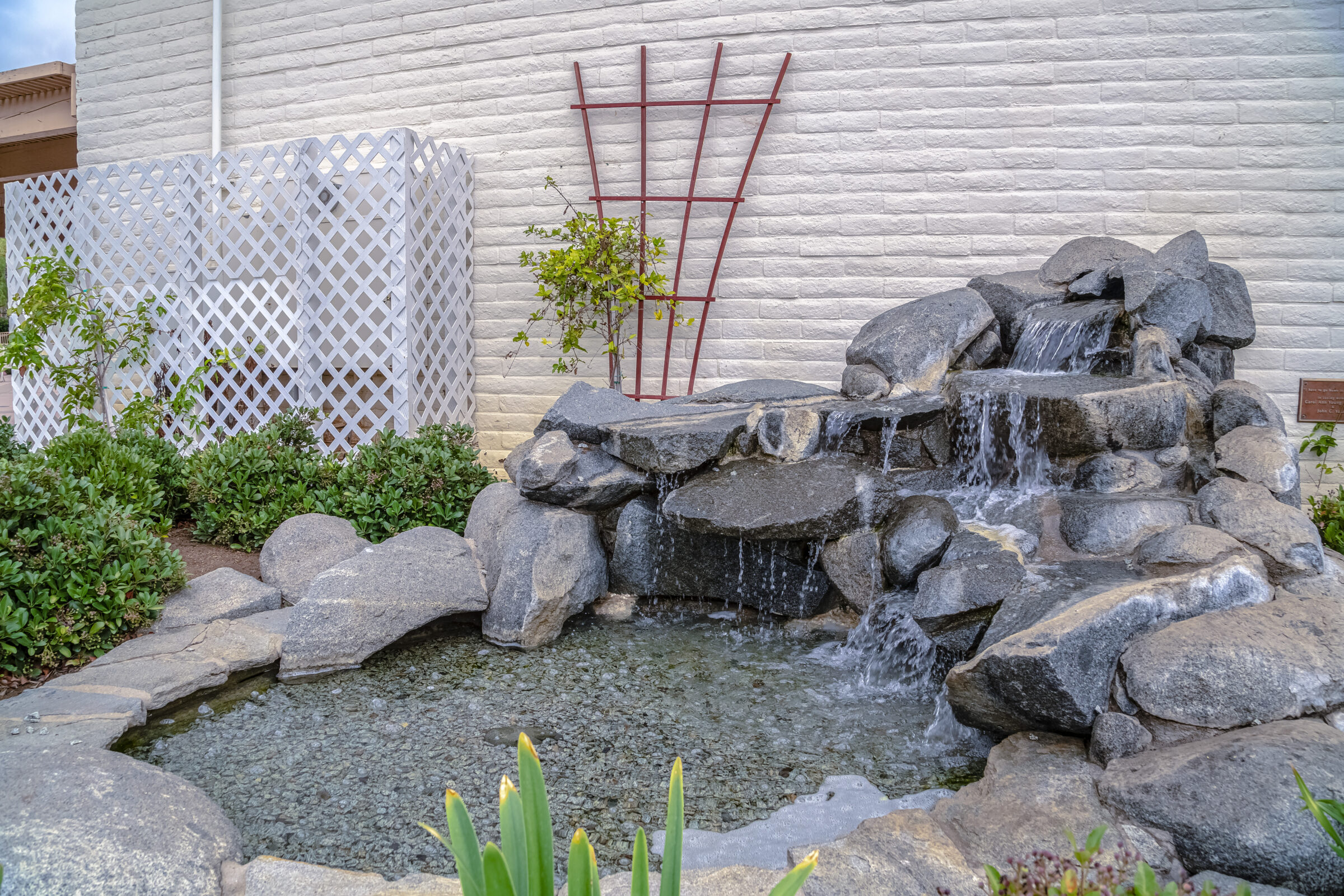 Rock waterfall and small pond beside a white lattice fence and brick wall, surrounded by greenery and plant vines.