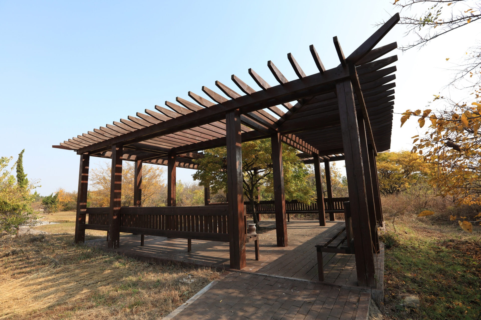 A wooden pergola stands in a scenic park with autumn trees, surrounded by grass and a clear sky.
