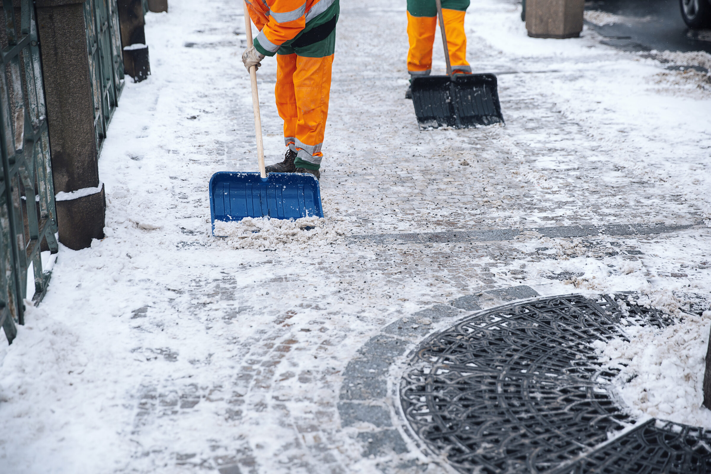 Two people in safety gear shovel snow from a city sidewalk, clearing a path on a cold winter day.