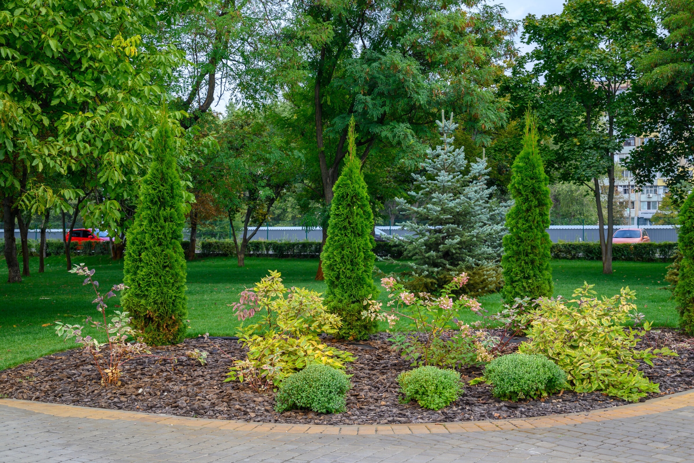 A tranquil park scene showcasing lush greenery, neatly mulched garden beds, and various trees, bordered by a stone path and distant buildings.