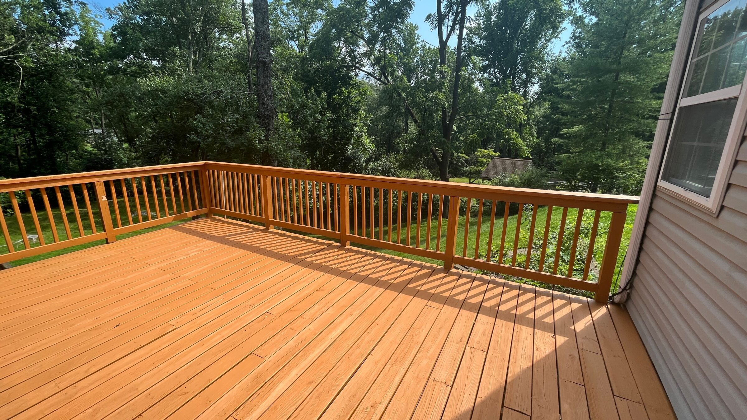 A wooden deck overlooking a lush, green backyard with tall trees, adjacent to a house featuring beige siding and white-framed windows.