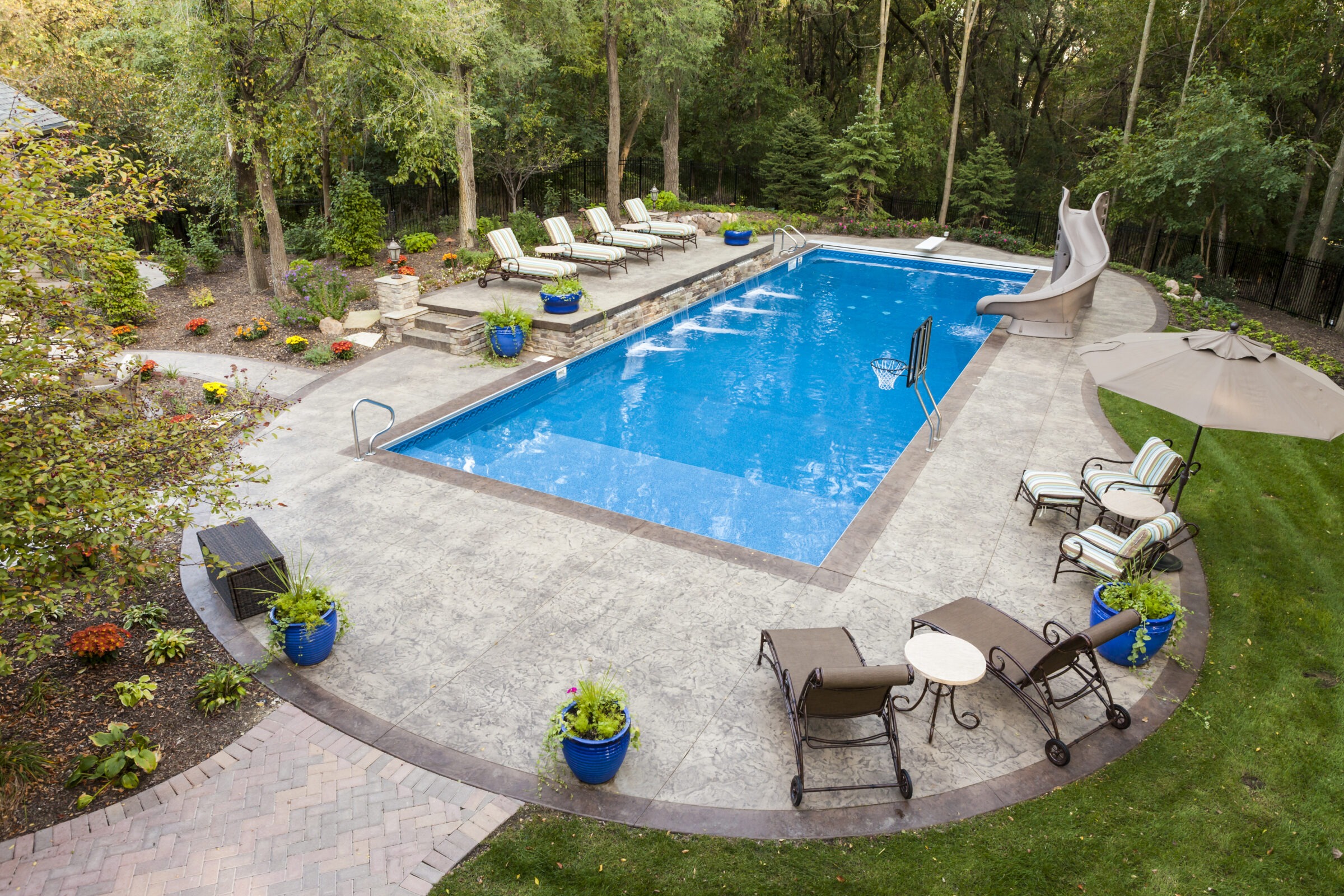 A backyard pool with lounge chairs, slide, and lush greenery. Potted plants and an umbrella add comfort and style to the setting.