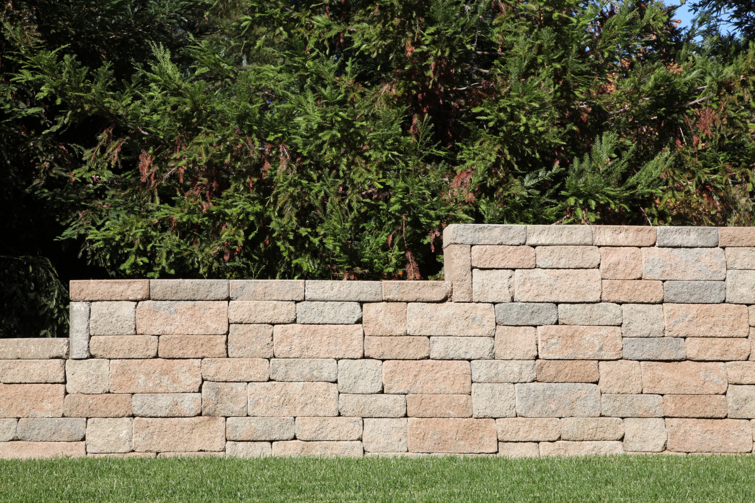 A stone retaining wall in a garden setting with evergreen trees in the background and well-maintained green grass in the foreground.