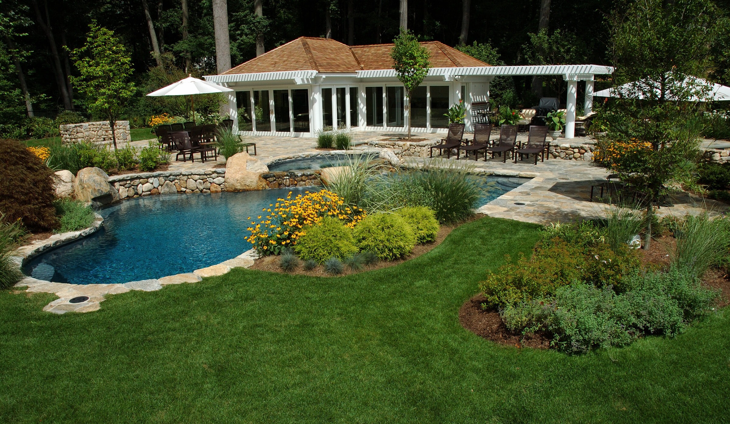 Luxurious backyard features a swimming pool, lush landscaping, stone patio with seating, and a cozy pool house surrounded by trees and greenery.