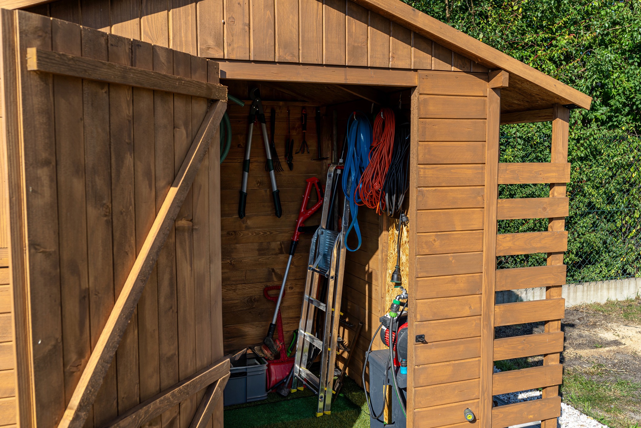 Wooden garden shed with open door, revealing tools, cords, and a ladder inside. Green foliage in the background.