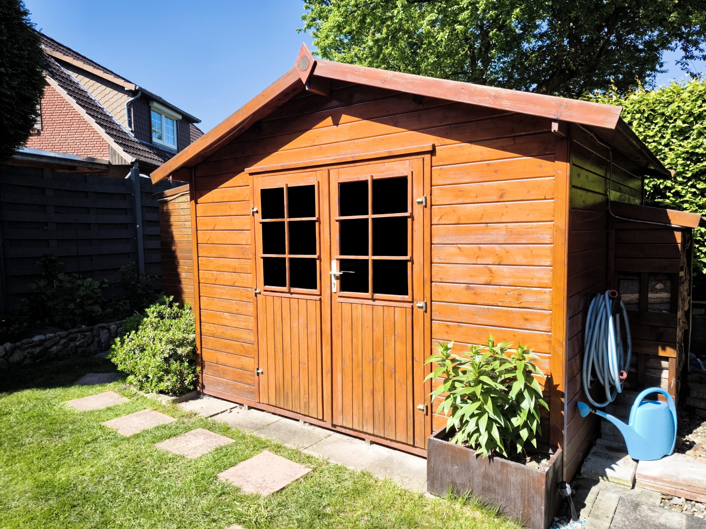 A wooden garden shed with double doors stands next to a house. A blue watering can and hose are nearby in the sunlight.