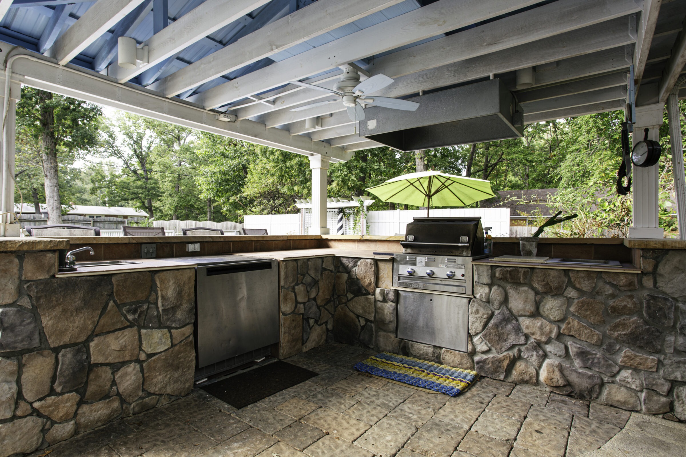 Outdoor kitchen with stone countertops, grill, and sink under a blue-roofed canopy. Green umbrella and trees visible in the background.