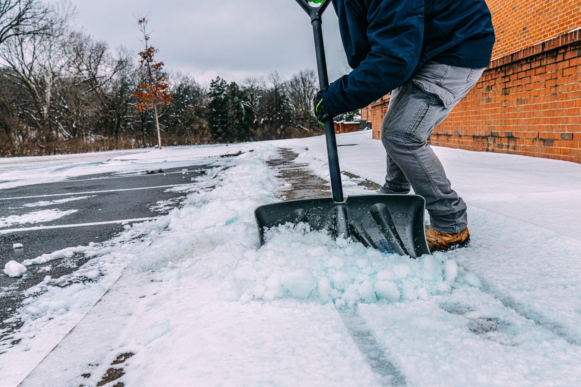 A person shovels snow on a path beside a brick building, surrounded by trees during wintertime. Overcast sky suggests a cold, wintry day.