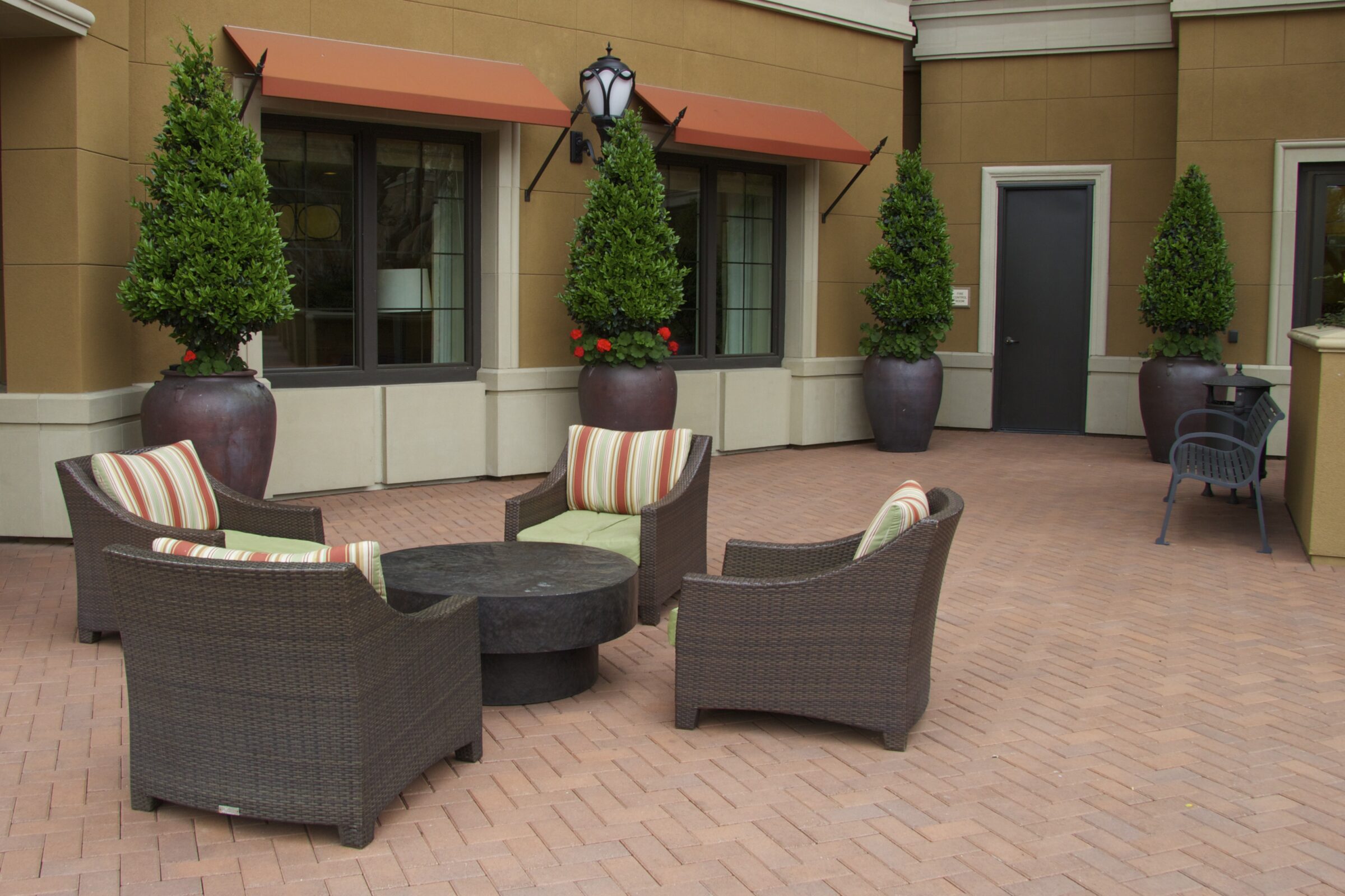 Outdoor seating area in a courtyard with wicker chairs, striped cushions, potted plants, and decorative lighting against a beige building facade.